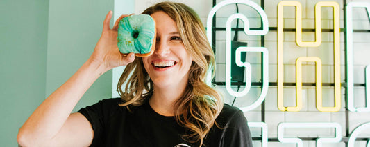 A female holding a blue glazed donut in front of her eye in a donut shop.