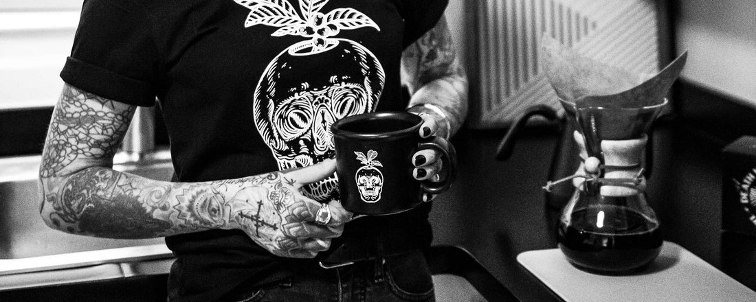 A female with tattoo sleeves holding a black mug of coffee next to a Chemex.