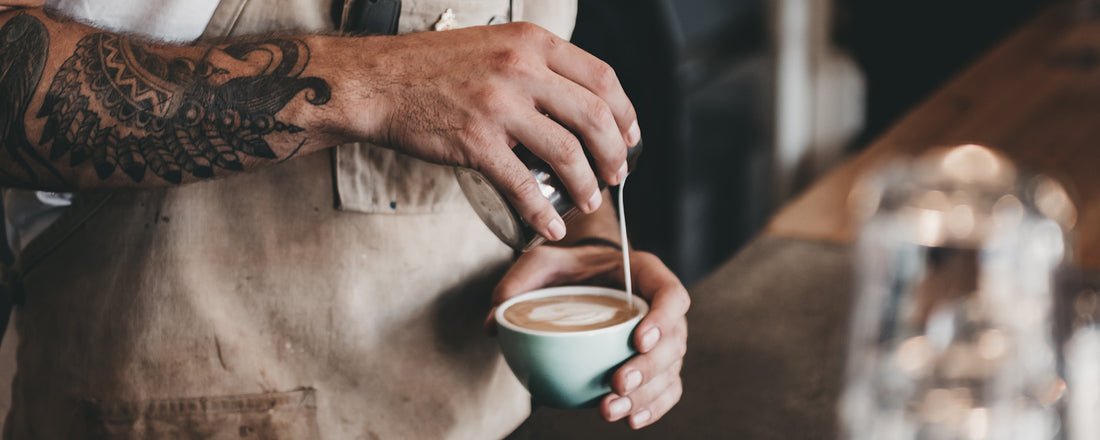 A man with a tattoo sleeve pouring coffee creamer into a mug of coffee. Photo Credit: Brent Gorwin via Unsplash.