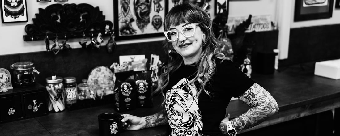 A woman with tattooed arms holding a mug and smiling in her tattoo shop.