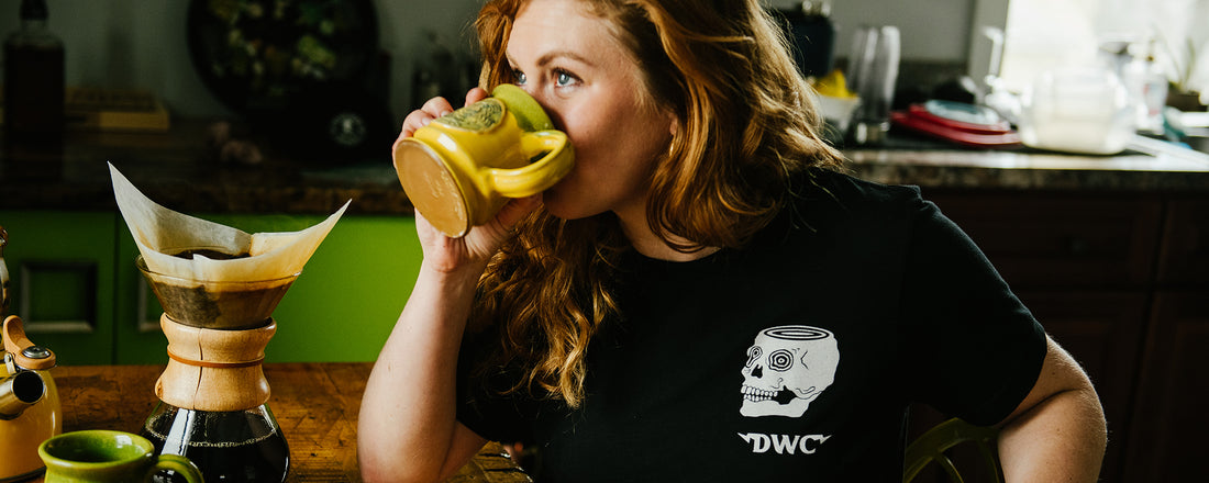 A woman drinking pour over coffee out of a yellow mug with a Chemex on the kitchen table.