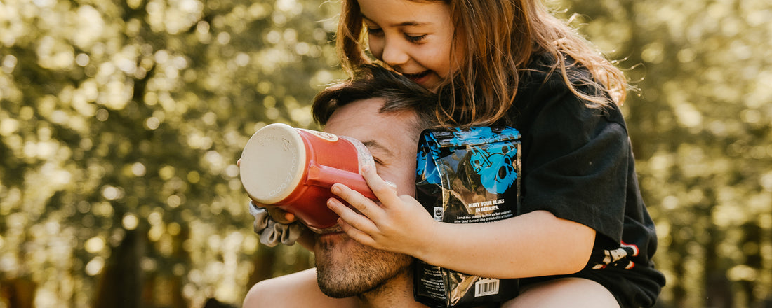 A father with his daughter on his shoulders pouring a mug of coffee into his mouth.