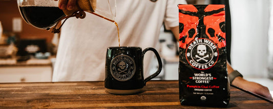 A Chemex pouring coffee into a black glazed Death Wish Coffee mug that is sitting next to a black and orange bag of Pumpkin Chai coffee.