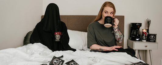 A female sipping coffee in bed next to the Grim Reaper holding a mug of coffee in bed.