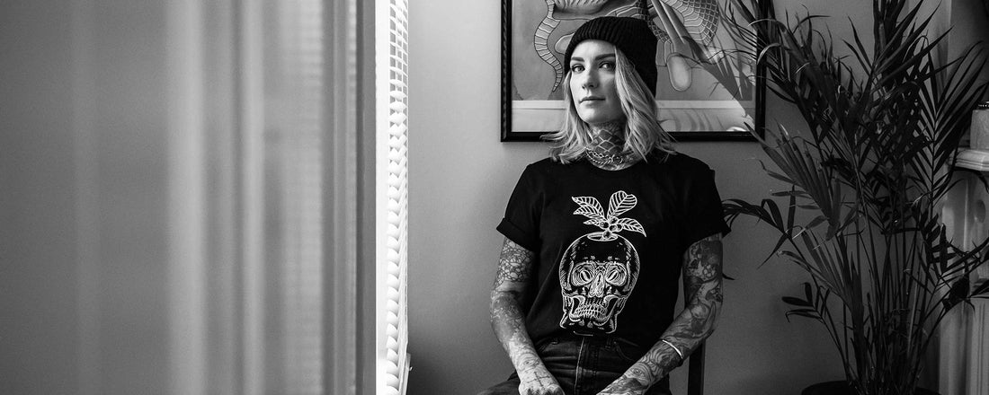 A woman with a sleeve of tattoos holding a mug of coffee with a skull and a coffee plant blooming out of it.