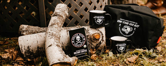 A campground scene featuring a pile of birch trees on the leafy ground with instant coffee, two mugs and a camping bag. 