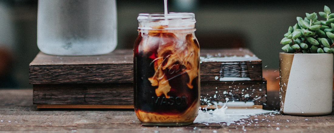 A mason jar of iced coffee with a plant-based coffee creamer being poured in. Photo Credit: Tyler Nix via Unsplash.
