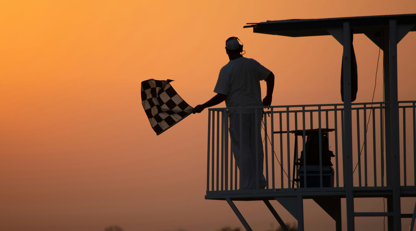 The silhouette of a man waving a checkered flag at sunset during a race. [Image Credit: Glen Rushton via Unsplash]